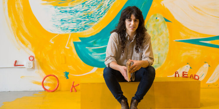 Photo of LK James sitting against a yellow mural by the artist.