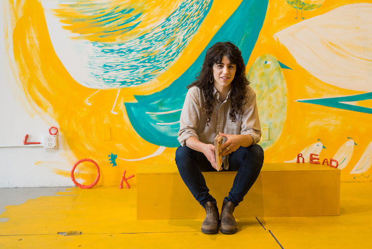 Photo of LK James sitting against a yellow mural by the artist.