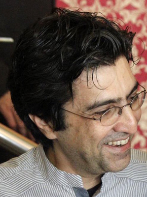 Photo of poet Kaveh Bassiri smiling with short black hair and glasses in front of a blurred background.