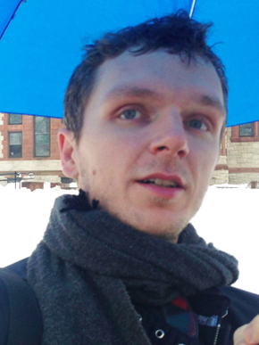 Mark Leidner, a light-skinned man with short hair, looks up at the camera while wearing a scarf and jacket and holding a blue umbrella over his head on a snowy day.