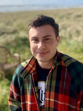 Photo of writer Milo R. Muise, a white transmasculine person in a plaid coat, standing in a green field with the ocean in the background.