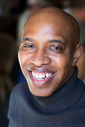 The photo of André Le Mont Wilson shows a middle-aged, bald Black man standing before a blurred background and wearing a black turtleneck sweater, a broad smile, and sparkly eyes.