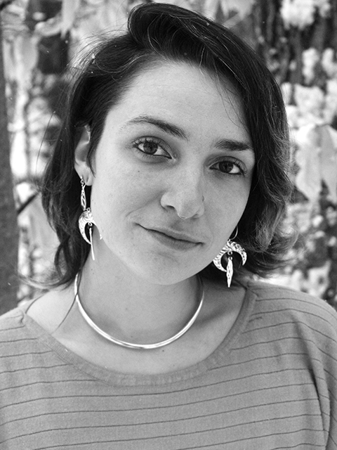 Black and white photo of an AFAB human with long dark hair, the sides of their head subtly shaved, outside in the forest with half-moon silver dangle earrings and the smallest sprinkle of snow in their hair.