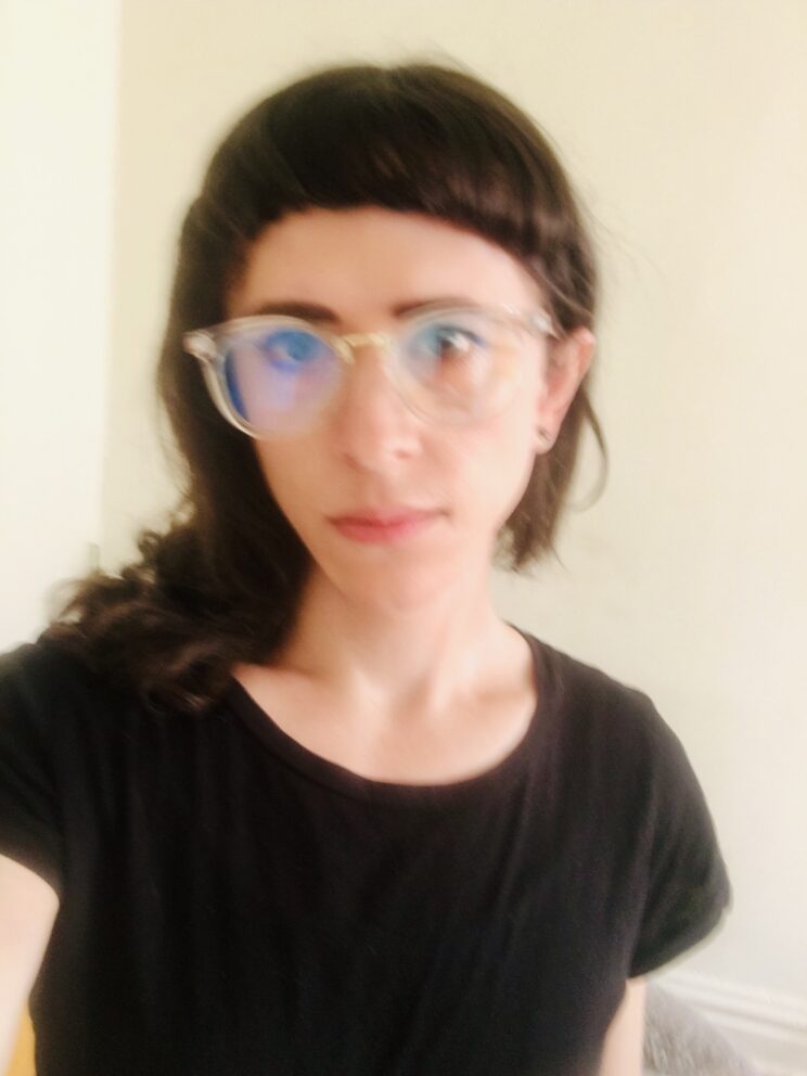 White trans woman with black bangs in black T-shirt and clear framed glasses against a corner of white apartment walls.
