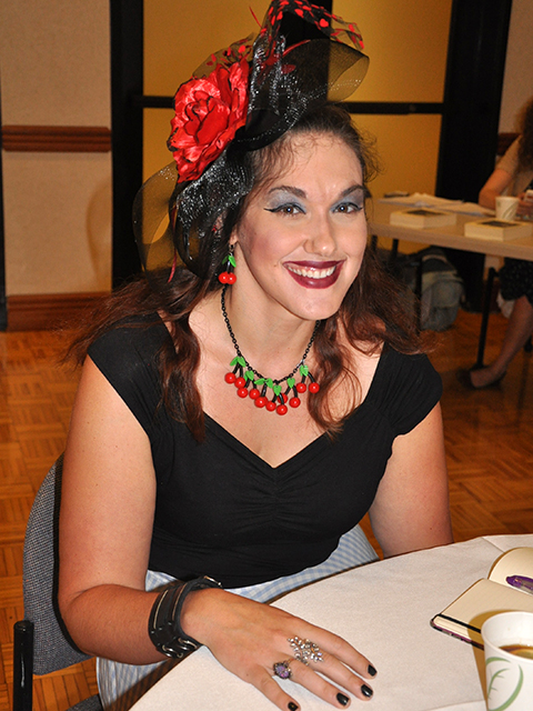 Head shot of Nicole Yurcaba, a white woman sitting at a table, wearing a black shirt and a black and red hat.