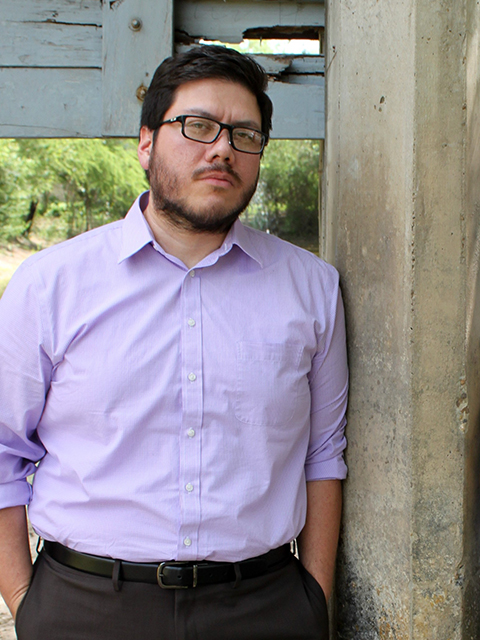 Photo of poet Rodney Gomezz, a bearded Mexican American, wearing black glasses and a light purple button-down shirt facing camera with hands in his slacks leaning against a wooden fence post.