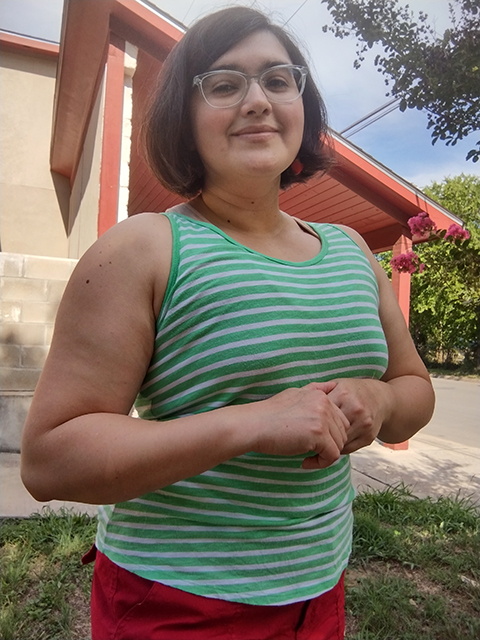 Person with chin-length, dark brown hair stands with elbows bent, hands clasped under their chest, wearing a green and white horizontal-striped tank top, red shorts, and strawberry earrings.