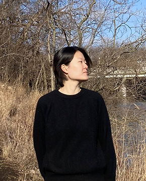 Light-skinned East Asian person with black hair in a black sweater standing in front of tall grass and trees.